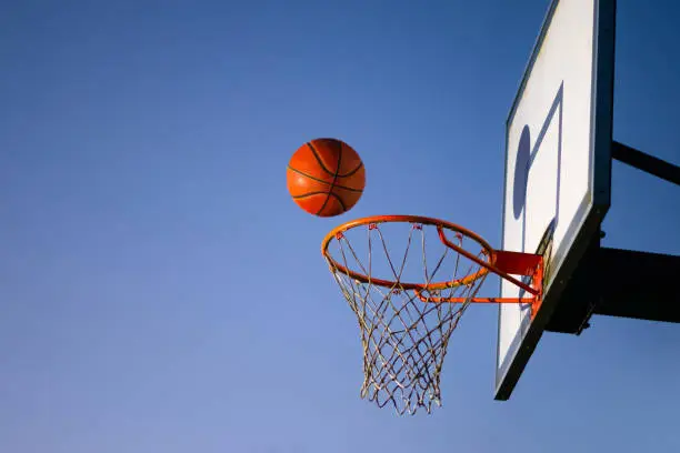 Photo of Street basketball ball falling into the hoop. Close up of orange ball above the hoop net with blue sky in the background. Concept of success, scoring points and winning. Copy space