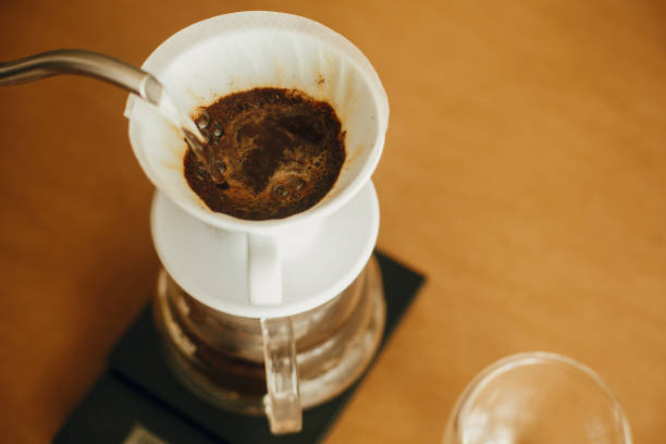 pouring hot water from steel kettle in filter with ground coffee in pour over on scale. alternative coffee brewing v60 closeup. barista making filter coffee on brown background - pouring coffee liquid coffee bean imagens e fotografias de stock