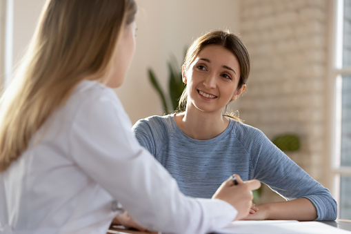Smiling millennial woman visit female doctor consulting about health complains, happy satisfied young client talk discuss treatment prescription with GP or physician at appointment in hospital