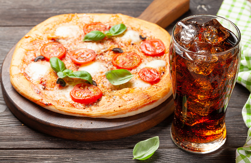 Glass of cola with ice and tasty homemade pizza with garden tomatoes, cheese and basil