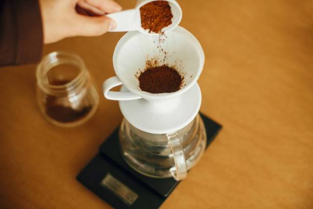 istock Hands pouring grounded coffee in filter pour over on glass kettle on scale. Preparing for alternative coffee brewing v60. Person holding spoon with grind coffee on brown background 1224220742