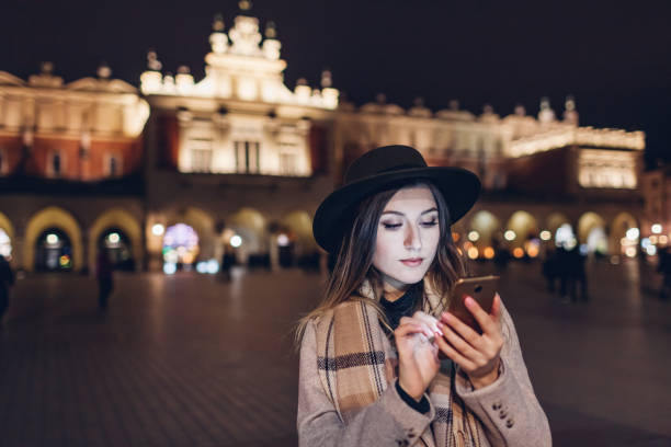 femme de touriste utilisant le smartphone la nuit sur la place de marché à cracovie pologne. fille vérifiant les médias sociaux d’information. voyage - krakow people poland church photos et images de collection