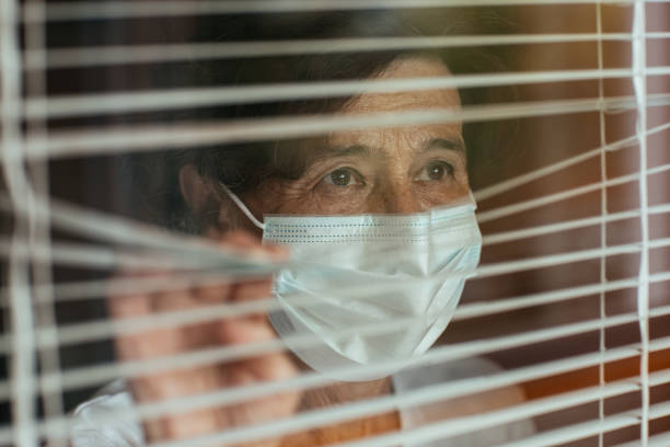 Close up of anxious old woman surgical mask looking through window blinds during self-quarantine. stock photo