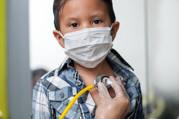 Child wearing a medical mask to prevent spread of virus is getting a heart screening with stethoscope placed on chest. stock photo