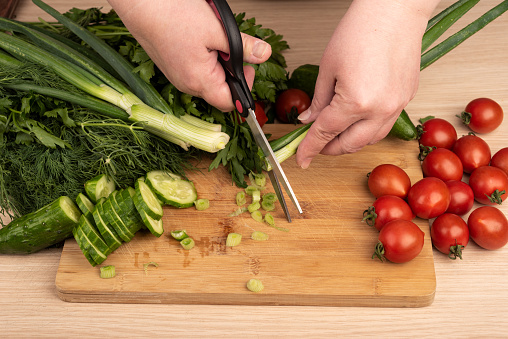 Woman cuts green onion with scissors over a cutting board. Greens, tomatoes and cucumbers on a wooden table. Rustic style.