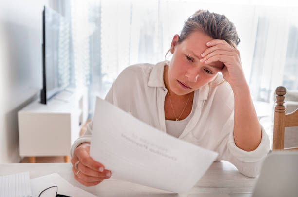 Woman looking worried holding paperwork at home. Woman looking worried holding paperwork at home. She is reading a financial bill or a letter with bad news. She looks very stressed and upset. There is a laptop computer on the table eviction photos stock pictures, royalty-free photos & images