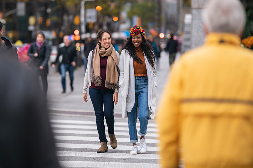 A multi-ethnic pair of female friends is exploring the city while on vacation. One of the women is Eurasian and the other is black. The beautiful and adventurous women are smiling. They are crossing a busy street surrounded by other pedestrians. It is a cool Autumn day and the women are wearing casual clothing.