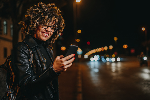 Cheerful young woman in night out using phone on city street