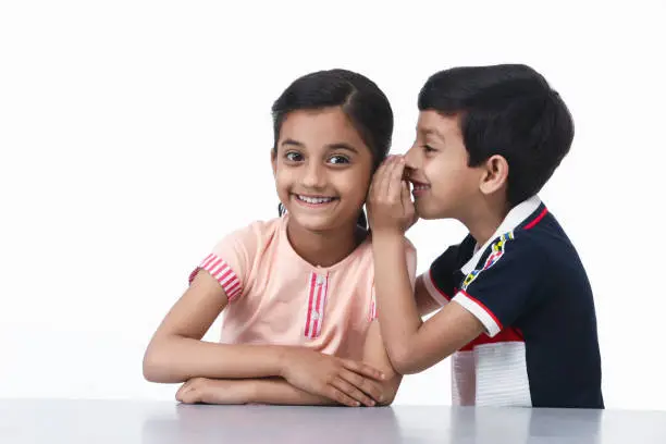 Elementary age cheerful Indian children sitting indoor & whispering something in the ears.