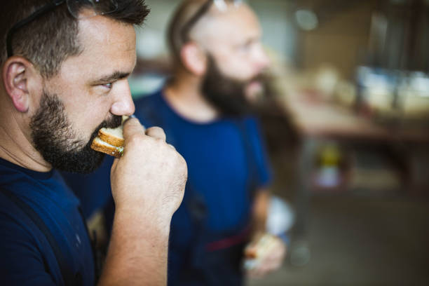 Manual worker eating sandwich on a break in a workshop. Profile view of young manual worker eating a sandwich on a lunch break in a workshop. His colleague is in the background. construction lunch break stock pictures, royalty-free photos & images