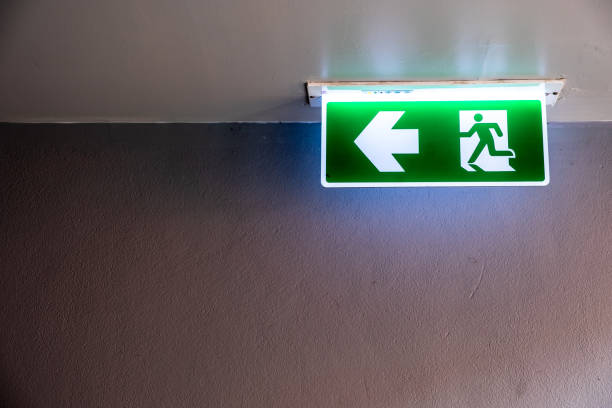 istock Fire exit sign with icon and arrow pointing to the left on green background hanging from the ceiling. 1224319063
