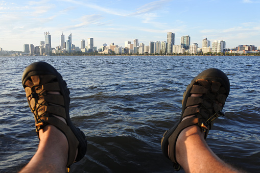 POV (point of view) of a person sitting on Swan River riverbank looking at the landscape view of  Perth city financial centre skyline at sunset in Western Australia.