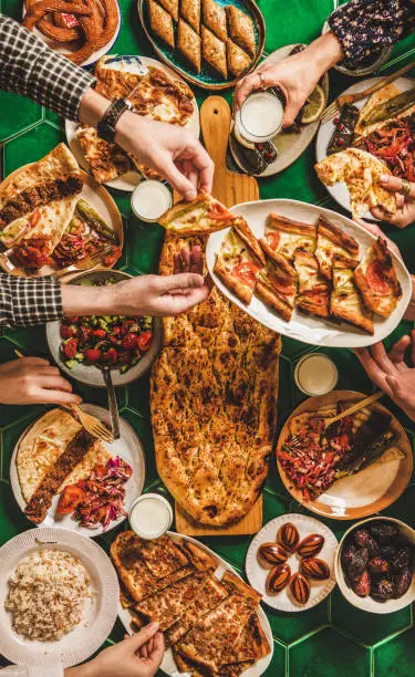 Muslim Ramadan iftar family dinner. Flat-lay of people feasting over table with Middle East food. Dates, kebab, flatbread, pide, borek, sweet, salad, top view. Ramazan fasting Turkish cuisine