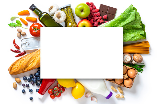 Top view of a large group of groceries like canned food, fruits, vegetables, eggs, a milk bottle, berries, mushrooms, nuts, pasta, a chocolate bar and bread against white background. At the center of the image is a white cardboard with copy space.