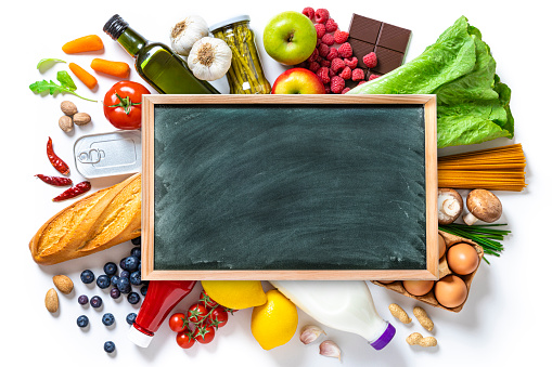 Top view of a large group of groceries like canned food, fruits, vegetables, eggs, a milk bottle, berries, mushrooms, nuts, pasta, a chocolate bar and bread against white background. At the center of the image is a blackboard with copy space.