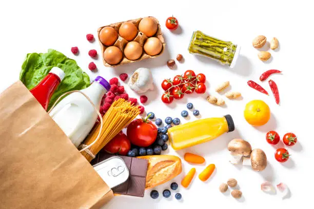 Top view of a paper bag full of canned food, fruits, vegetables, eggs, a milk bottle, berries, mushrooms, nuts, pasta, a chocolate bar and bread. The paper bag is laying on the white background and the food is coming out from it.