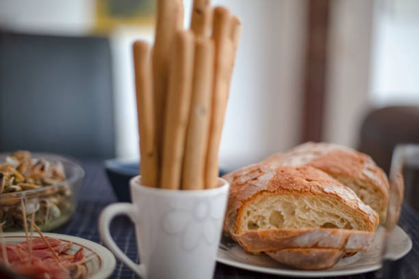 View of table prepared for brunch, with sliced rustic bread, cup with toasted breadsticks and slices of ham...