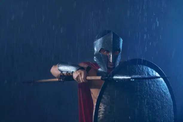 Portrait of spartan in armor and red cloak fighting outdoors with spear. Front view of man in helmet hiding behind iron shield and pointing weapon in bad cloudy rainy weather. Ancient sparta concept.