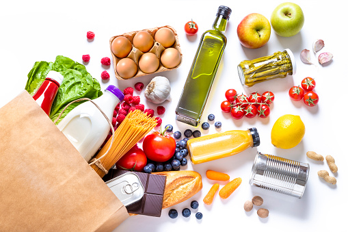 Top view of a paper bag full of canned food, fruits, vegetables, eggs, a milk bottle, berries, mushrooms, nuts, pasta, a chocolate bar and bread. The paper bag is laying on the white background and the food is coming out from it.
