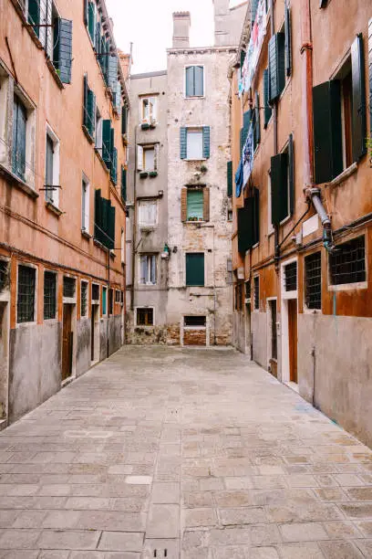Photo of Close-ups of building facades in Venice, Italy. Dead end Venetian street. Five-story houses with blue-green wooden shutters on the windows, linen is dried in the windows, an old stone floor.