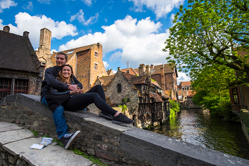 A couple of tourists pose for a photo on a picturesque bridge next to a typical canal in the city of Bruges.
