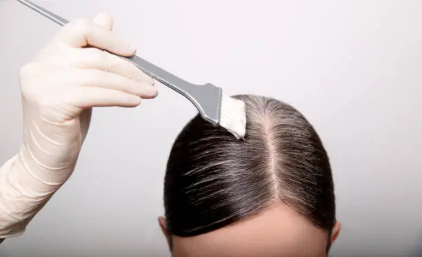 Photo of Woman dyeing gray hair root with brush and hair color, close up