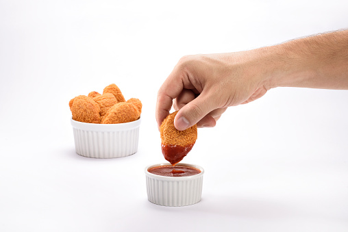 A hand dipping a delicious and crispy orange chicken nuggets on a ketchup sauce, isolated on white background, with a portion of nuggets served on a ramekin on the background. Ideal to illustrate a restaurant, snack at home, or to insert a different background for any other purposes.