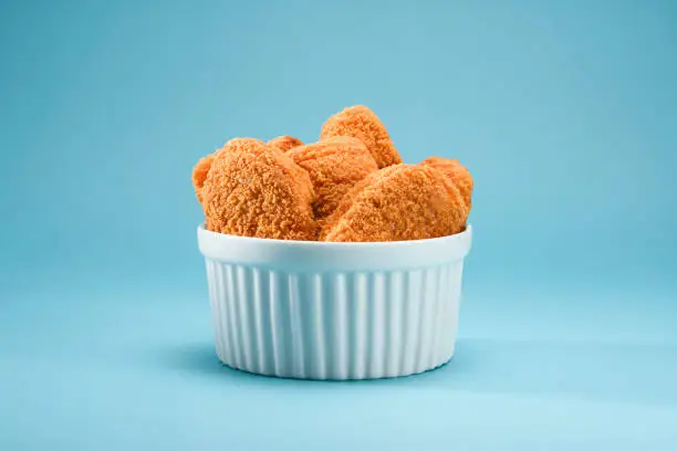 A portion of delicious and crispy chicken nuggets on a white ramekin, on a teal or blue background. A popular fast food  made from chicken meat that is breaded or battered, then deep-fried or baked. Ideal to illustrate a restaurant or snack at home on a teal and orange theme.