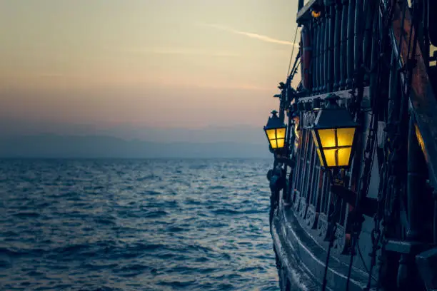 old wooden vintage pirate ship on sea water surface in sunset evening romantic time with yellow light from soft focus lantern in overboard space