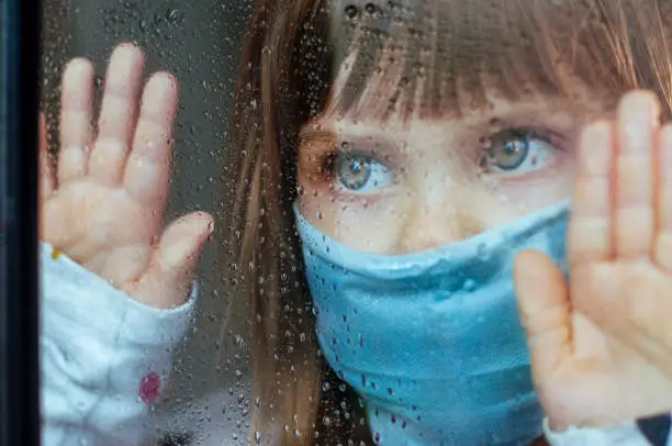 Little girl in face mask looking in the window glass with rain drops. Selective focus on the eyes. Social isolation stay at home during Pandemic COVID-19 concept.