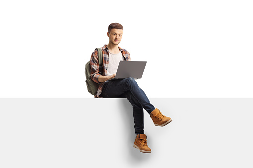 Male student with a laptop computer sitting on a blank panel and looking at the camera isolated on white background