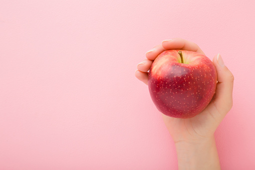 Young woman hand holding red apple on light pink table background. Fresh fruit. Pastel color. Closeup. Point of view shot. Empty place for text. Top down view.
