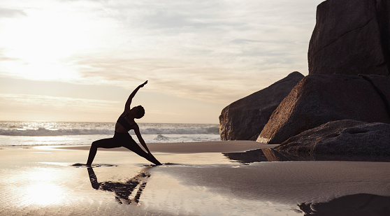 Full length shot of an athletic young woman stretching and practicing yoga on the beach at sunset