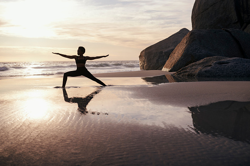 Full length shot of an athletic young woman stretching and practicing yoga on the beach at sunset