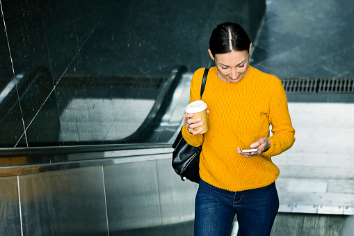 Woman holding documents and coffee cup and moving up on escalator