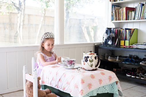 A beautiful little girl enjoys a tea party. Her teddy bear is sitting in the chair next to her.