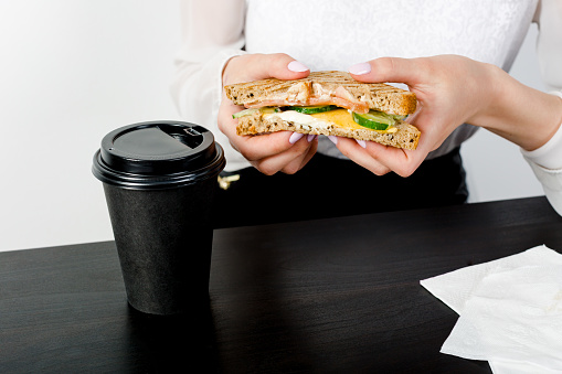 Women's hands holding a bite sandwich over a desk with cop of coffee on a white background. Unhealthy fast food at work place concept.