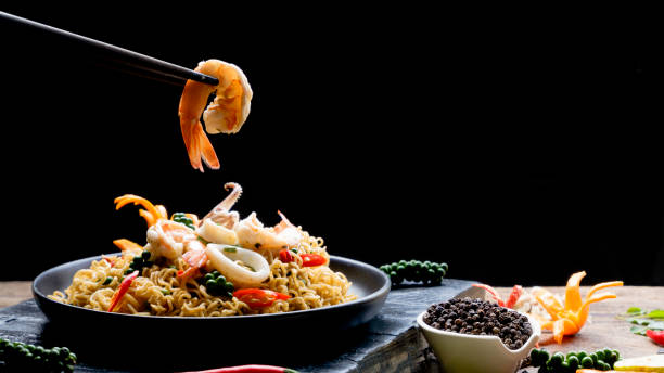 chopstick pickup shrimp with stir fry instant noodles spicy seafood in black plate with various spices on black slate in dark background Selective focus at chopstick picking up shrimp with stir fry instant noodles spicy seafood in black plate with various spices on black slate in dark background, Asian meal concept prawn seafood photos stock pictures, royalty-free photos & images