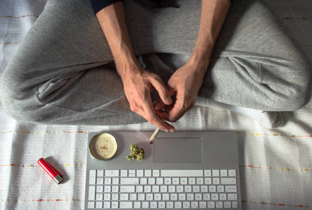 vista superior de uma pessoa sentada em uma cama fumando maconha enquanto usa um laptop para assistir a um vídeo ou filme. cannabis e conceito de tecnologia. - unrecognized person - fotografias e filmes do acervo
