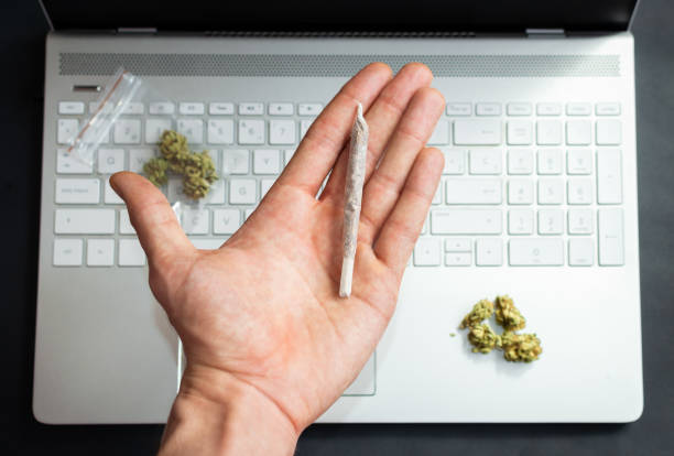 istock Top view of hand holding a big marijuana joint with computer and cannabis buds on background. Concept of marijuana and technology or office. 1224410537