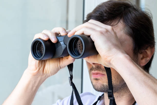 istock Handsome bearded man looking through binoculars at the window with a view of the neighboring house for observing neighbors, environment or nature during quarantine. Selective focus. Closeup view 1224412991