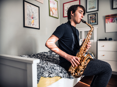 Teenage boy enjoying his hobby: He is  in his bedroom playing alto saxophone.