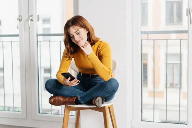 Young attractive woman in jeans and yellow sweater sitting on a chair with her legs crossed and looking at the screen of mobile phone with pretty smile. Front portrait with copy space