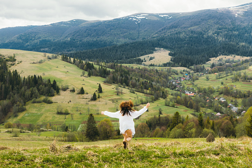 Beautiful young woman with curly hair, in ethnical old school white dress and hair accessory from the woods walking on the fresh green mountain hills with view of the snowcapped peaks in Carpathian Mountains, Ukraine during bright spring day - feeling freedom and happiness