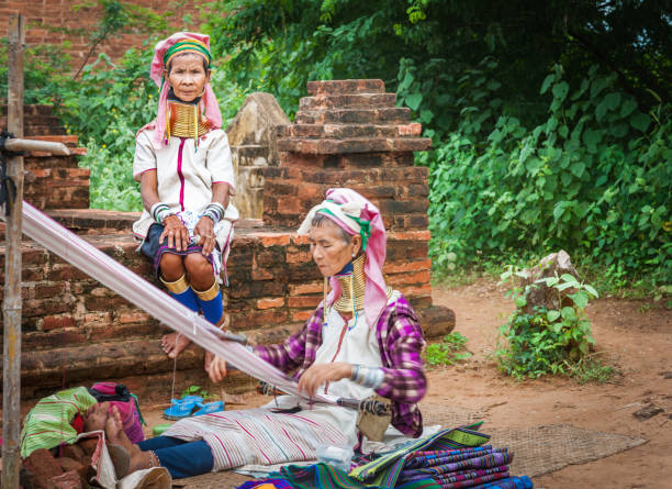 Myanmar Travel Images Kayan women with brass rings around necks Bagan Myanmar - October 29 2013; Two Kayan women with brass rings around necks one weaving while other in selective focus sitting wall looks on. kayan neck rings stock pictures, royalty-free photos & images
