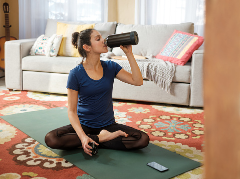 A healthy young latin woman sitting on mat in the living room and drinking water from a reusable bottle.