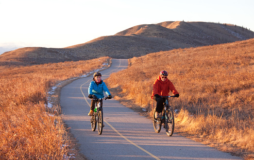 An evening image of two people biking down a rural pathway in the rolling river valley on the prairies. Image taken in rural Alberta near Calgary. Front view. Image taken in December.