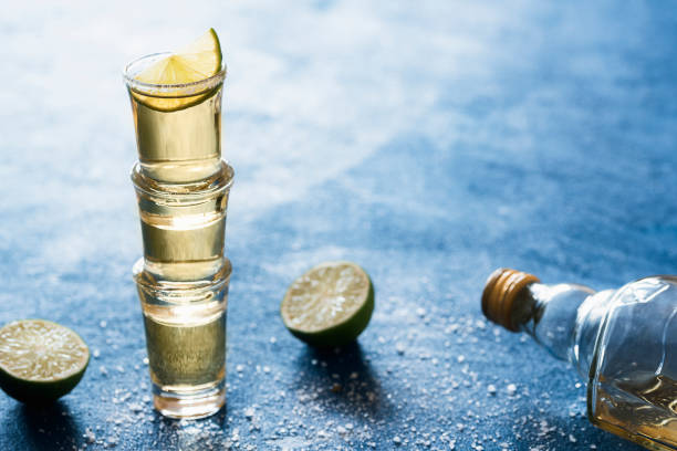 Golden tequila in shot glass with salt and lime, on a blue table, backlight. Three tequila shots stacked together. Mexican traditional beverage.