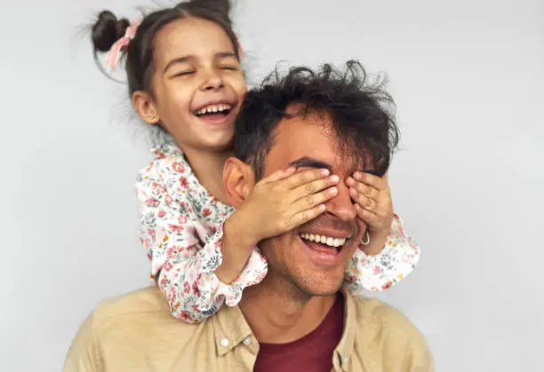 Closeup image of smiling little girl embraces her dad, shares love together. Handsome man playing with kid peekaboo game. Playful daughter cover the eyes with hands of her father for a surprise.