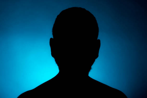 silhueta limpamente definida de uma pessoa masculina virada para a direita contra um fundo escuro com um holofote azul e área brilhante logo atrás do busto. - defined - fotografias e filmes do acervo